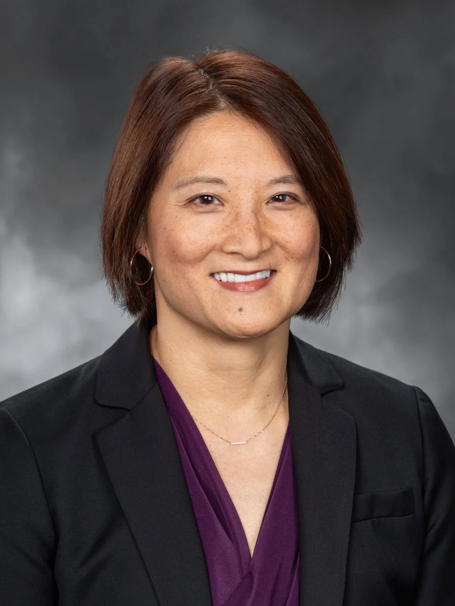 Professional headshot of a woman with short brown hair, wearing a black blazer and purple blouse, smiling against a gray background.