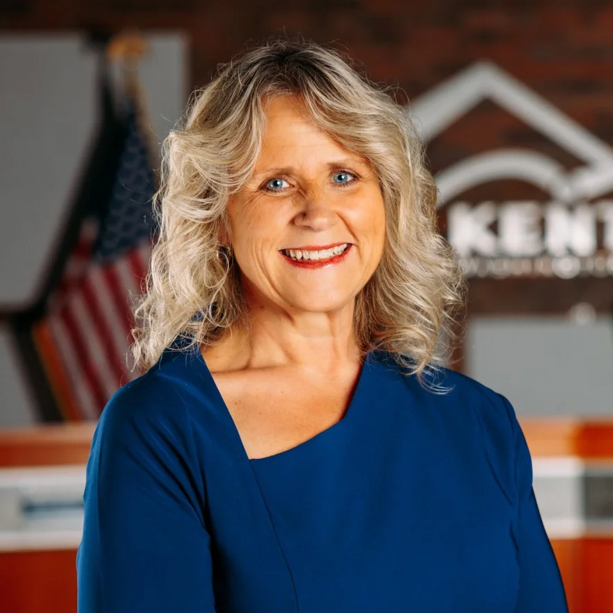 Smiling woman with curly blonde hair wearing a royal blue dress, standing indoors with American flag and a sign that says 'KENT' in the background.