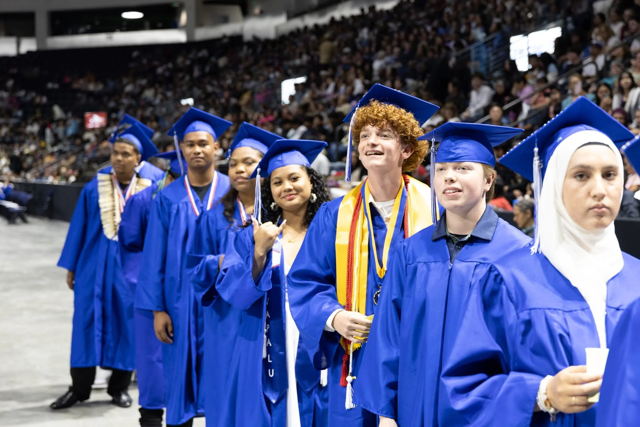 Graduates wearing blue caps and gowns at a ceremony, standing in a line with some holding hands or medals, in a large indoor arena filled with seated audience.