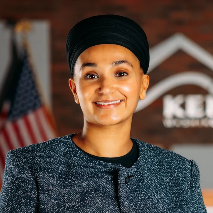 A woman with a headwrap smiling at the camera, with an American flag in the background and a sign that reads 'KELA'.
