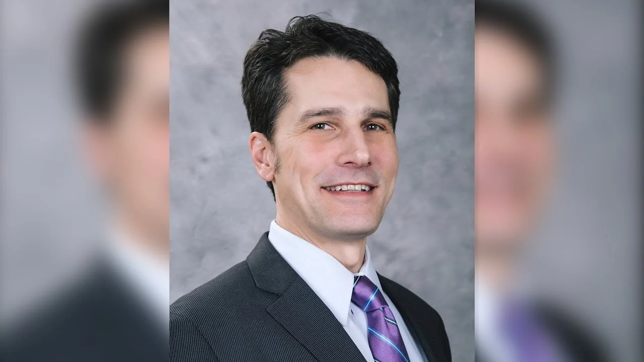 Headshot of a man with dark hair, wearing a dark suit, white shirt, and a purple striped tie, smiling against a grey textured background.