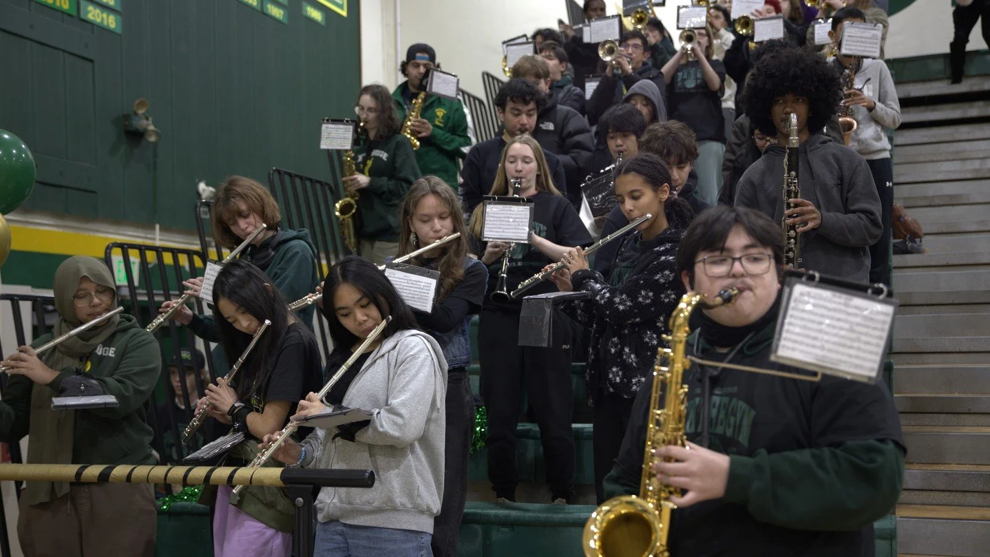 Group of students performing in a school band, playing various wind instruments including flutes, clarinets, saxophones, and saxophones, on a gymnasium staircase decorated with green and yellow balloons.