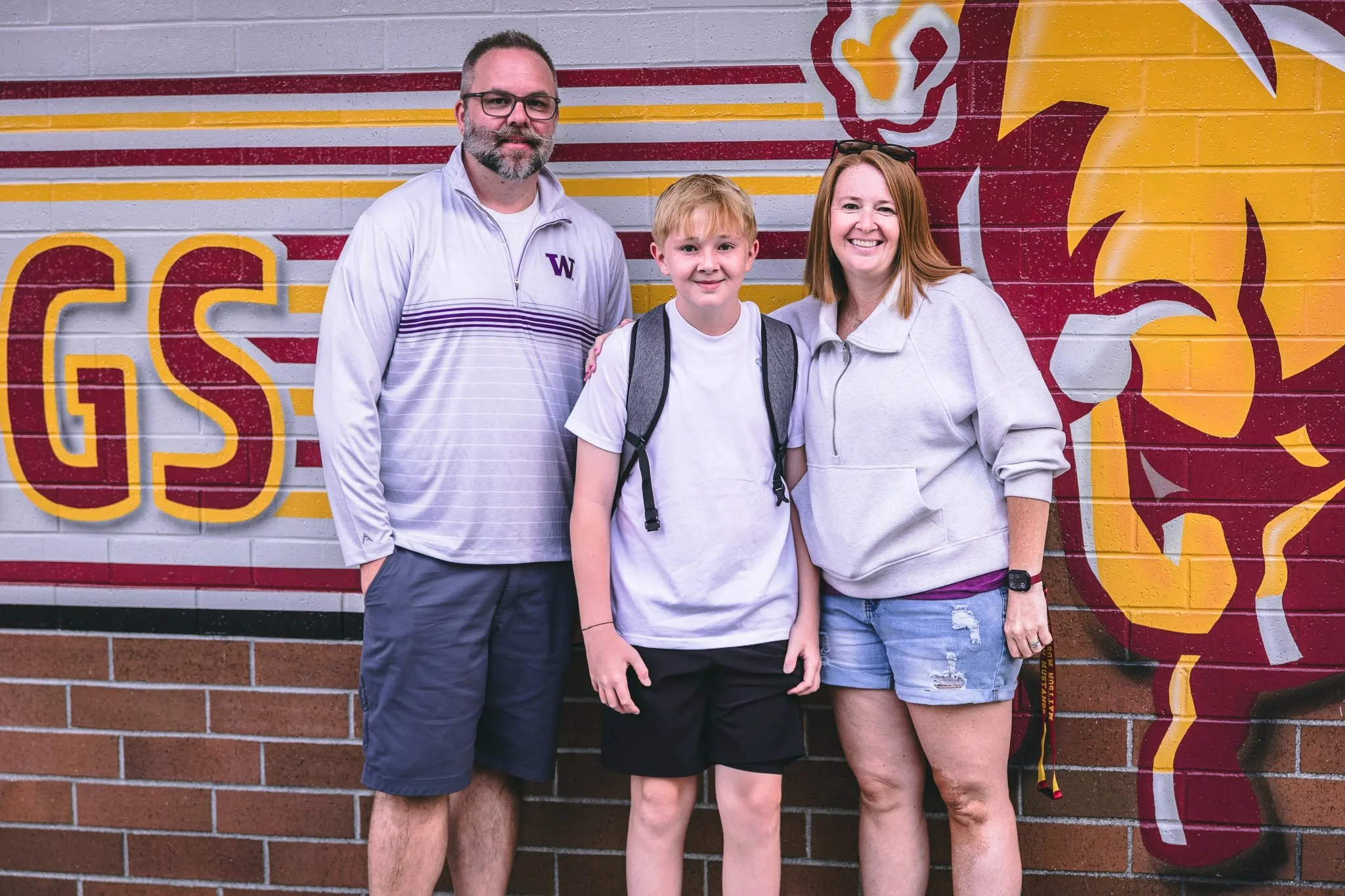 A family of three standing in front of a colorful wall mural at a school or sports event. The man has glasses, a beard, and is wearing a white jacket. The woman has red hair, sunglasses on her head, and is wearing a light-colored hoodie. The boy has blond hair, a white T-shirt, and a backpack.