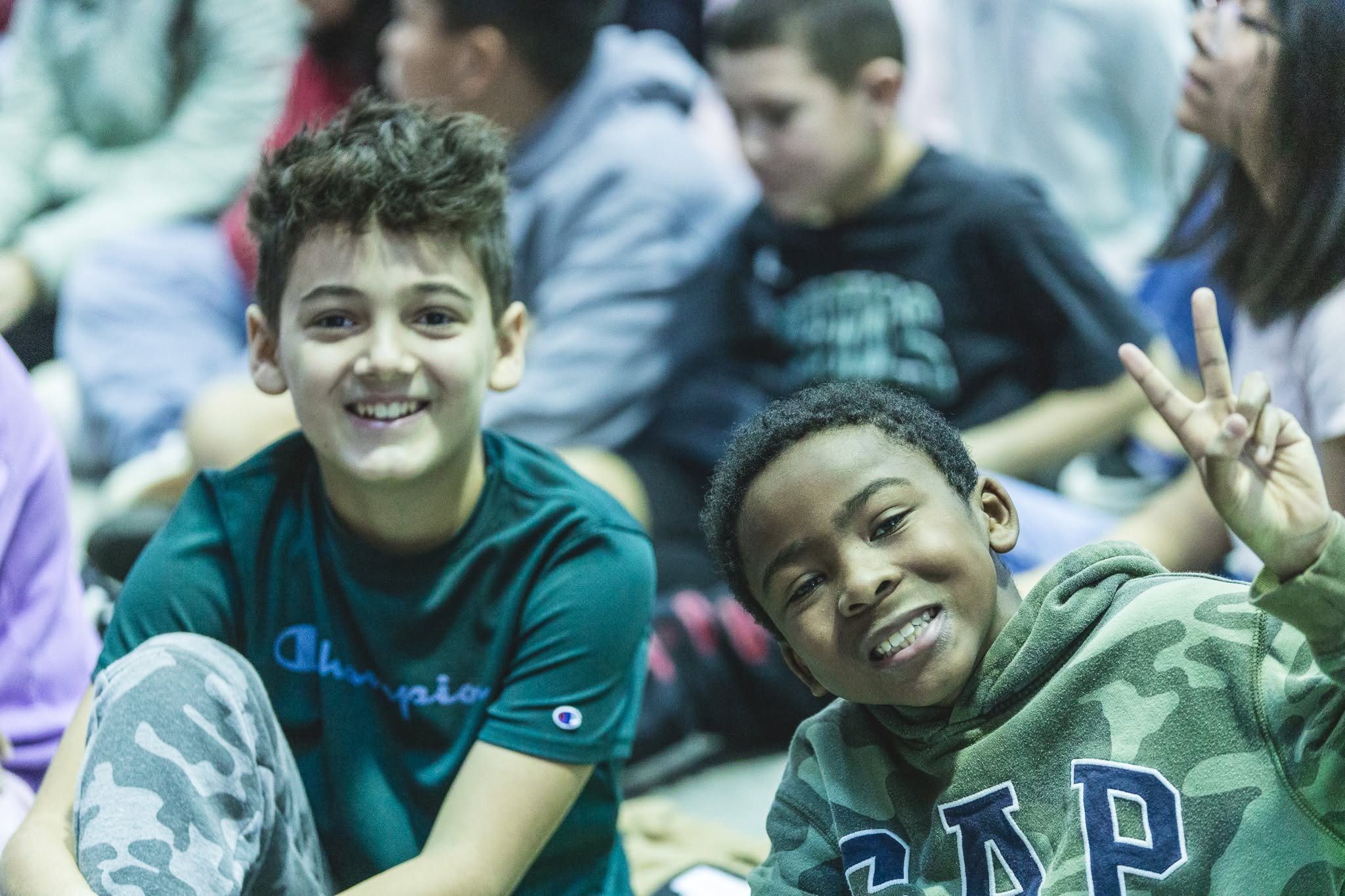 Group of smiling children and teenagers sitting together, some making peace signs, in an indoor setting.