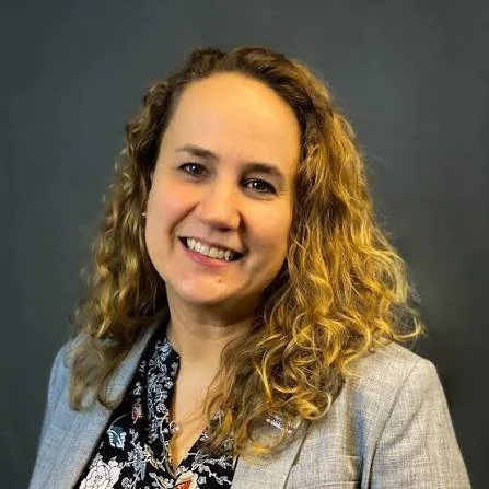 A woman with curly blonde hair smiling, wearing a light gray blazer over a floral blouse with a dark background.