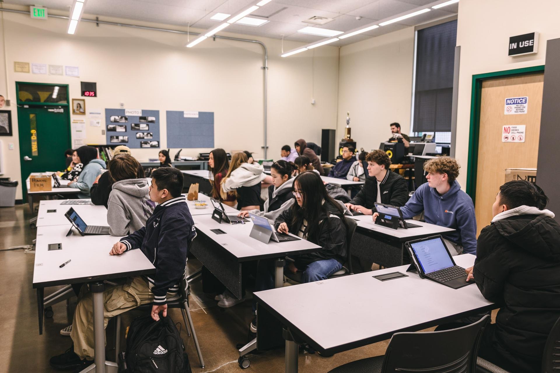 A classroom filled with students sitting at desks working on laptops. The room has beige walls, a large window with blinds, and educational posters on the walls. Students are focused and engaged, with some wearing hoodies and jackets.