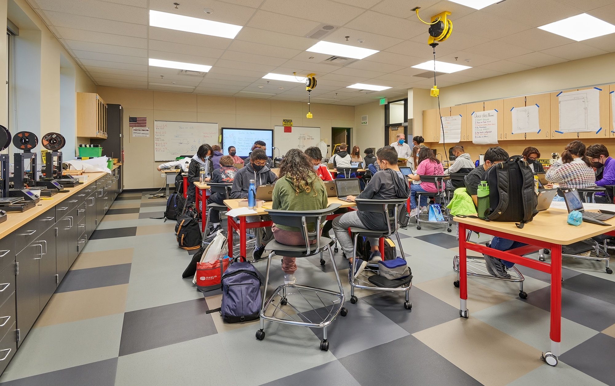 A classroom filled with students seated at red and black tables, working on laptops and reading books. The classroom has whiteboards, cabinets, and ceiling-mounted retractable cords. Students are wearing masks, and backpacks are placed beside or under their chairs.