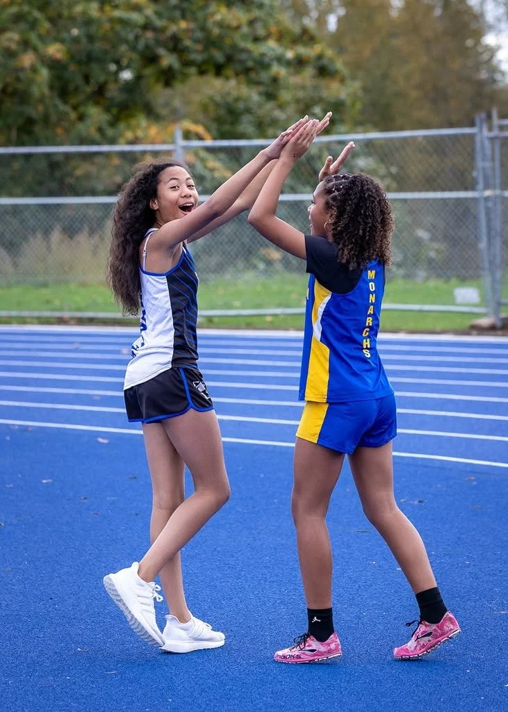 Two young girls on a blue athletic track, dressed in sports uniforms, are giving each other high fives and smiling.