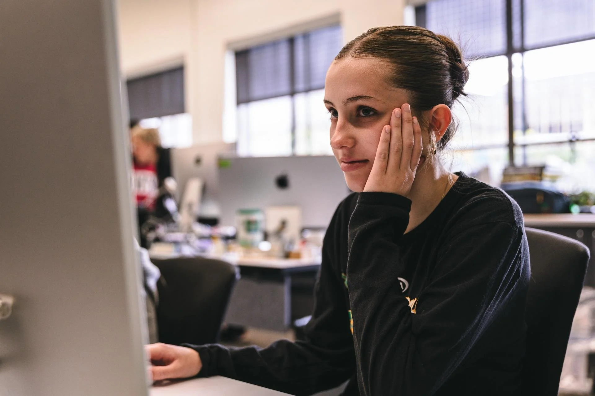 Young woman sitting at a desk in an office, touching her face and looking at a computer screen.