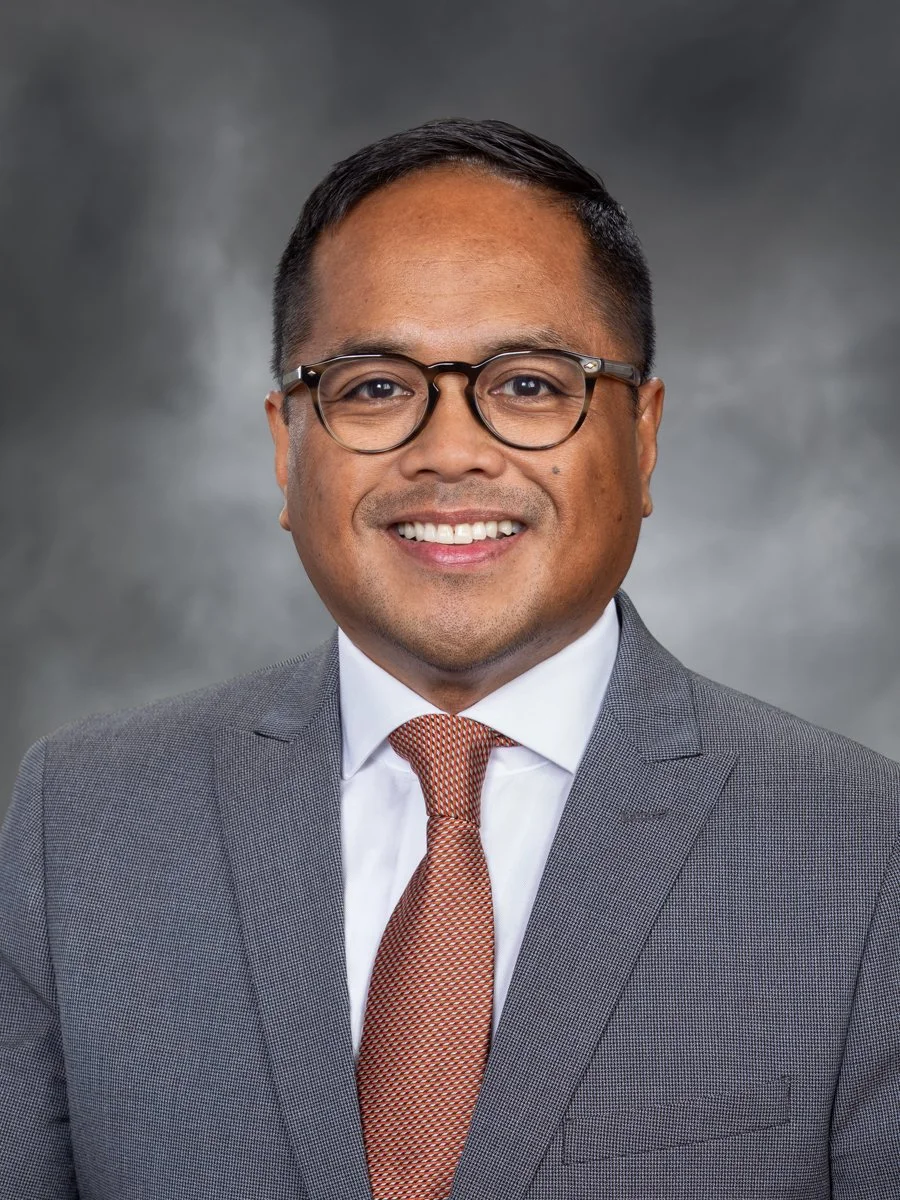 Professional headshot of a smiling man wearing glasses, a gray suit, white shirt, and orange tie against a gray background.