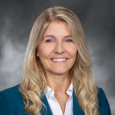 A woman with long blonde hair smiling, wearing a white shirt and a dark blazer against a dark gray background.