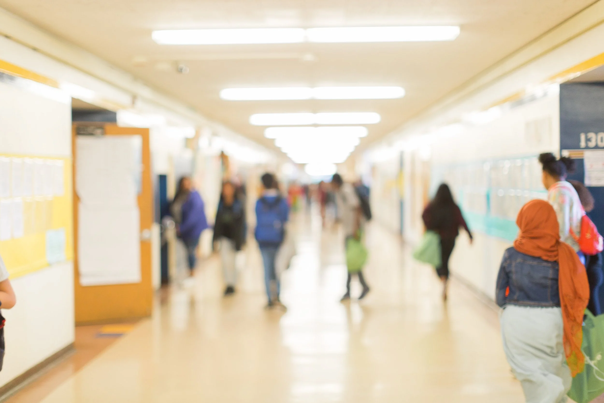 School hallway filled with students, lockers, and brightly lit ceiling lights.