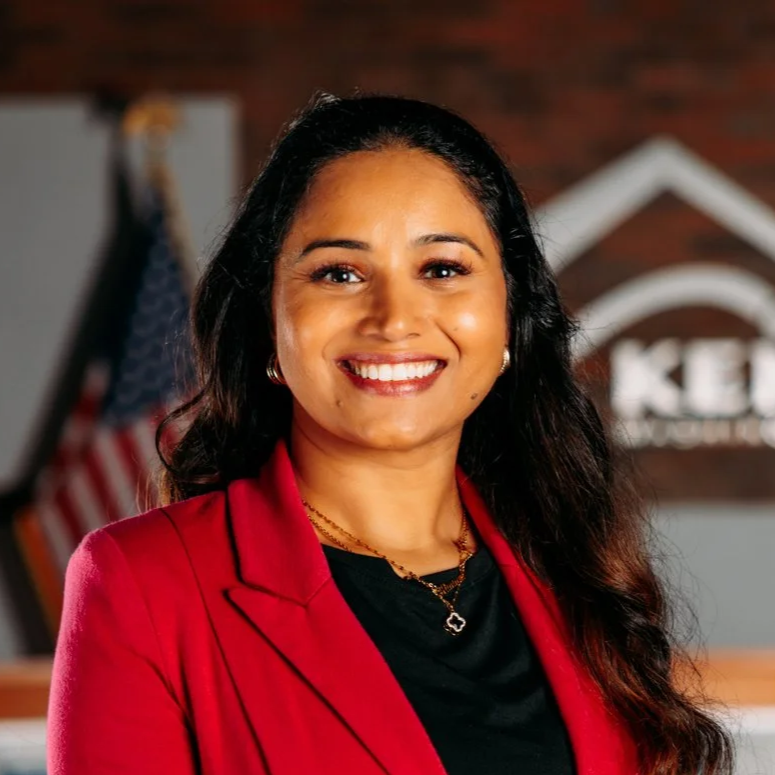 Smiling woman with long dark hair wearing a red blazer and black top, standing in front of a wall with a logo and an American flag.