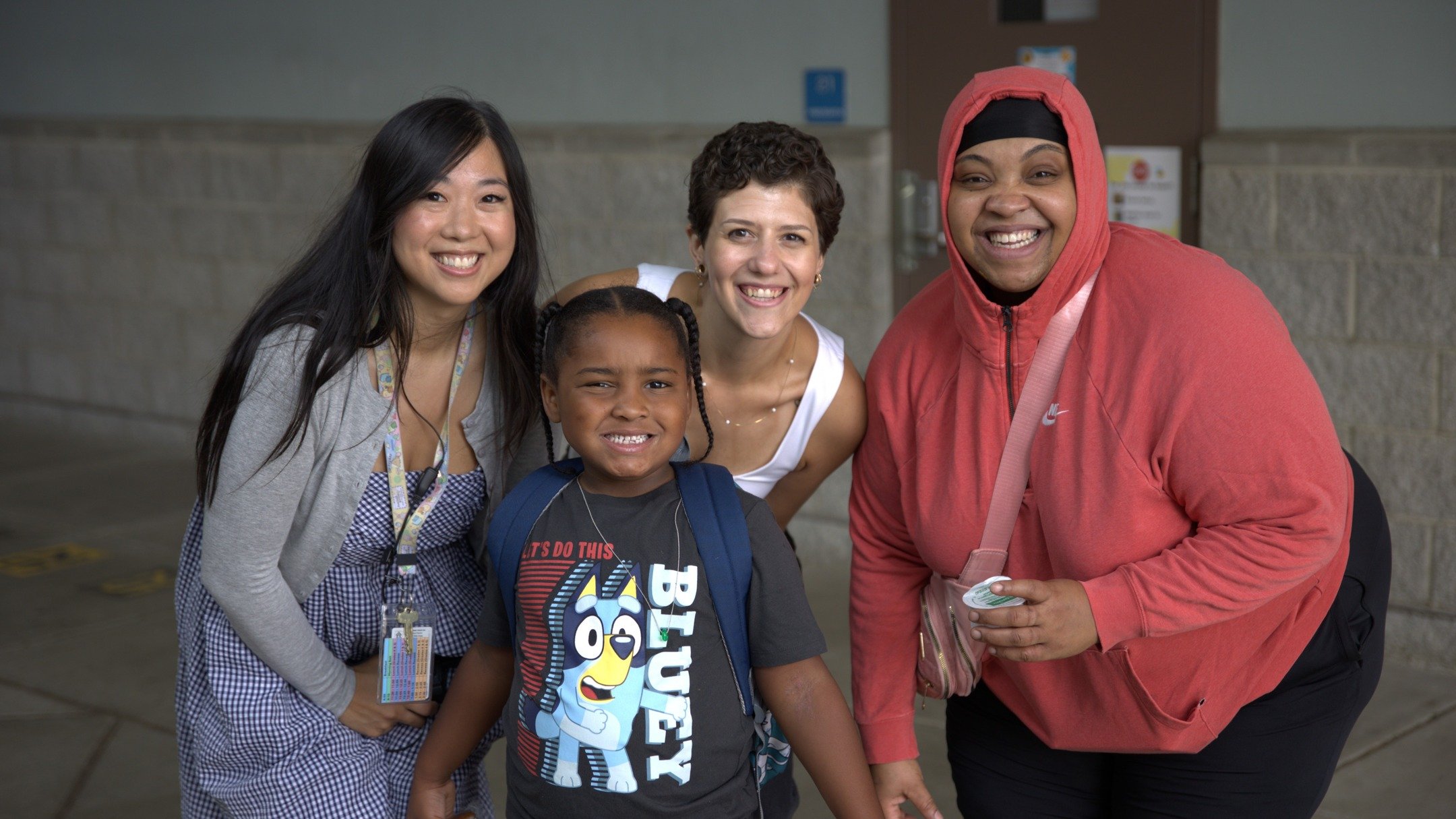 A group of five people, including a child, smiling and standing close together in an indoor setting, possibly a school or community center.