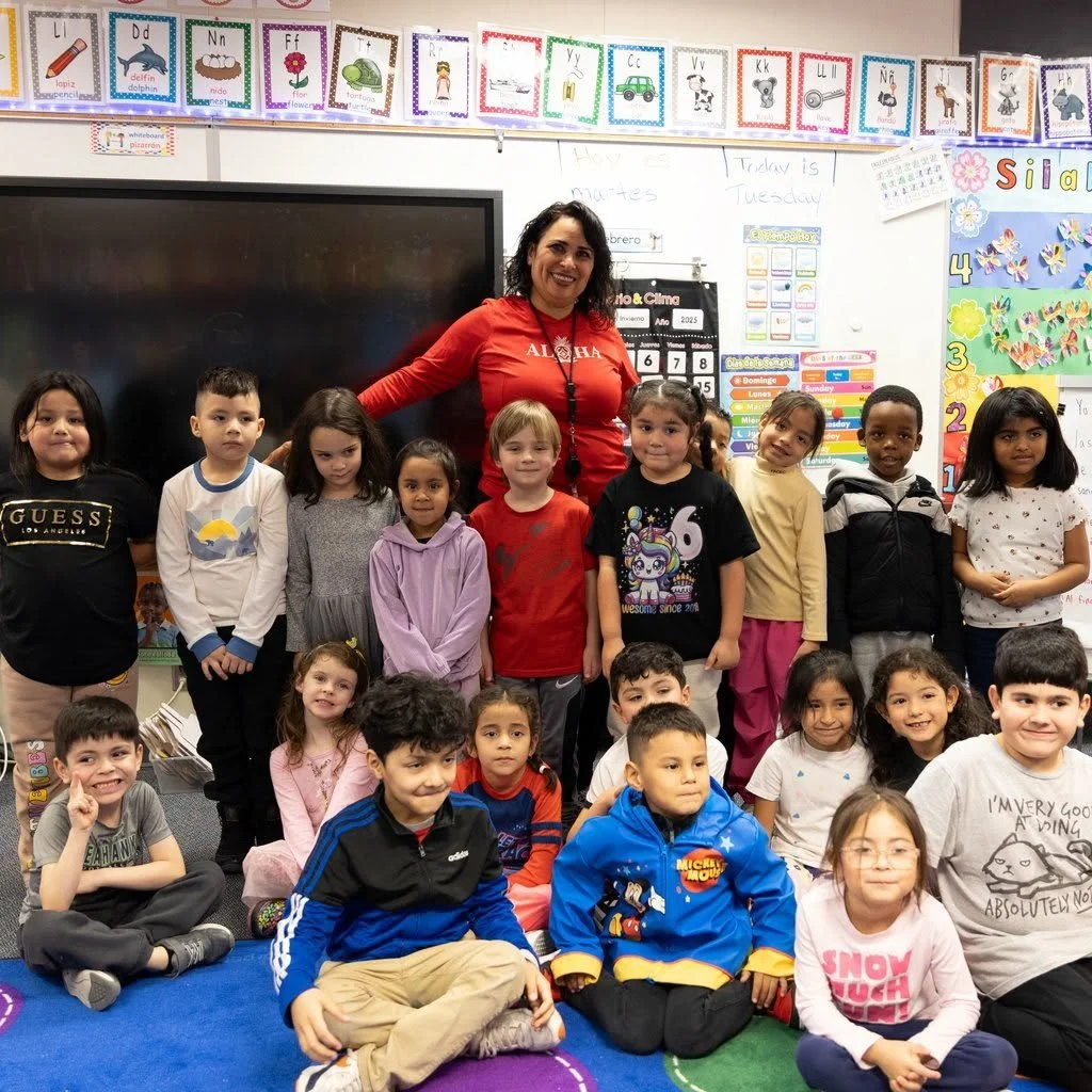 A group of young students and their teacher in a classroom, posing for a photo. The classroom has colorful educational posters, a whiteboard, and a large screen.