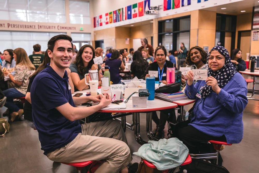 Group of diverse people sitting around a large table in a modern, well-lit conference room, attending an event or workshop, with flags hanging from the ceiling and motivational words on the wall.