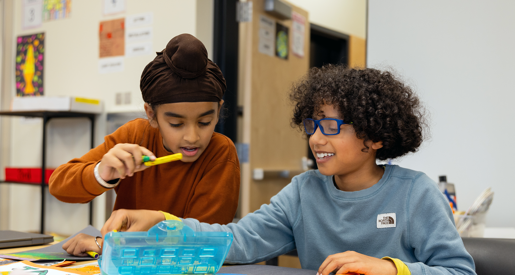 Two children, a girl with a brown headscarf and a boy with curly hair and glasses, are sitting at a table in a classroom. They are smiling and working on a project together, with various art supplies and papers on the table.