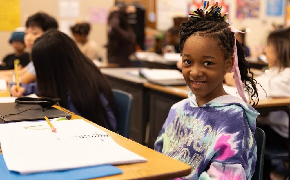 Smiling young girl with braided hair and a colorful hoodie sits at her desk in a classroom filled with students, with notebooks and writing utensils on her desk.