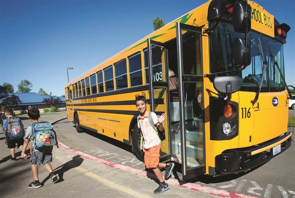 A group of children boarding a bright yellow school bus at a school parking lot on a clear sunny day.