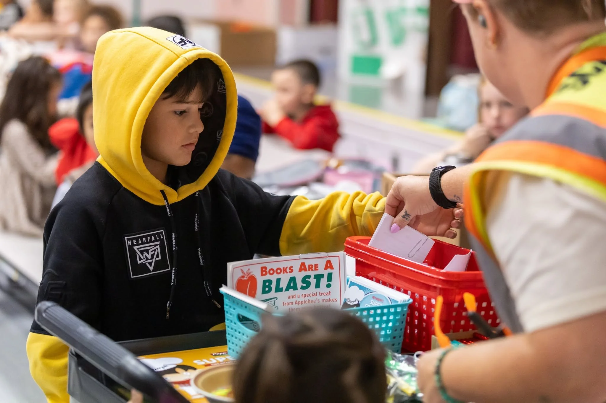 A young boy wearing a yellow hoodie and a black jacket is at a checkout counter in a store, looking at the cashier who is scanning items. There are other children in the background, and a colorful basket on the counter contains books and snacks. The environment suggests a busy, cheerful retail setting, likely a bookstore or grocery store, with a sign that says "Books Are A Blast!"