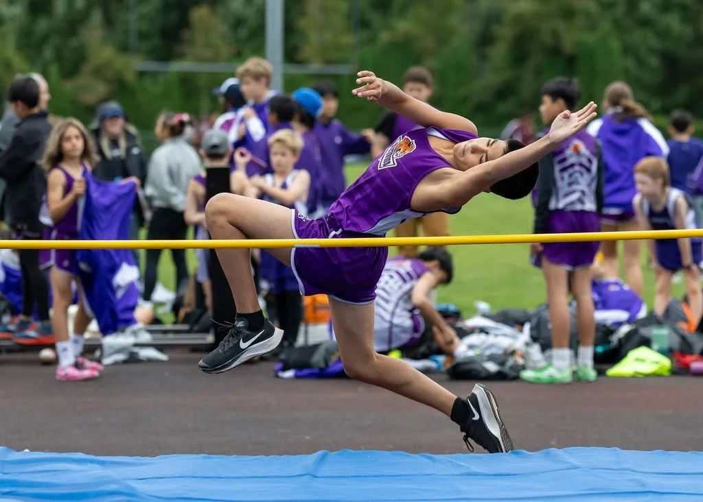Young female athlete in purple uniform competing in a long jump event, mid-air over the sandpit, with spectators and other athletes in the background.