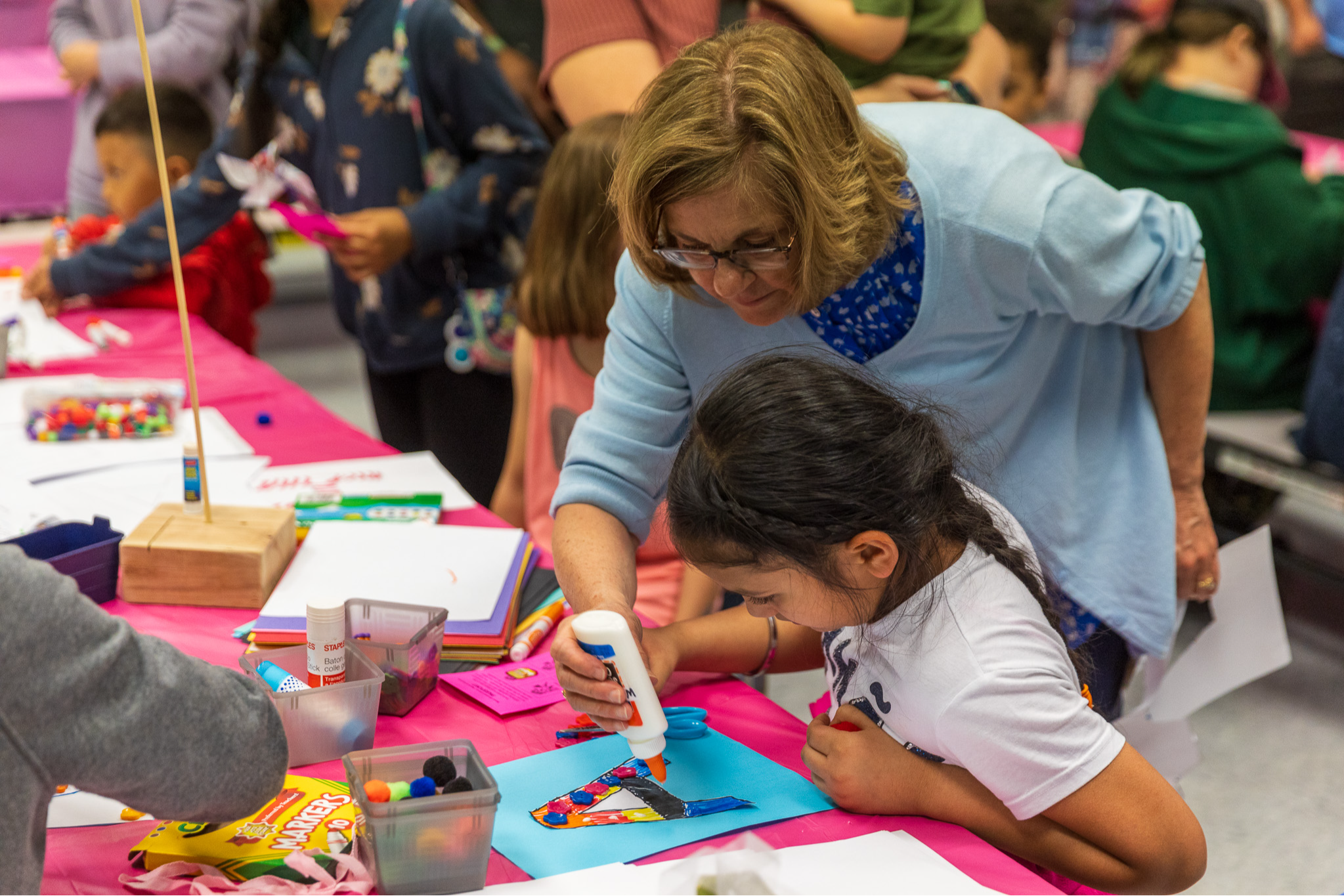 A woman with glasses and shoulder-length blond hair leaning over a young girl with dark hair, working on a craft project at a table covered with art supplies and colorful paper, in a busy, colorful room filled with children and adults engaged in arts and crafts activities.