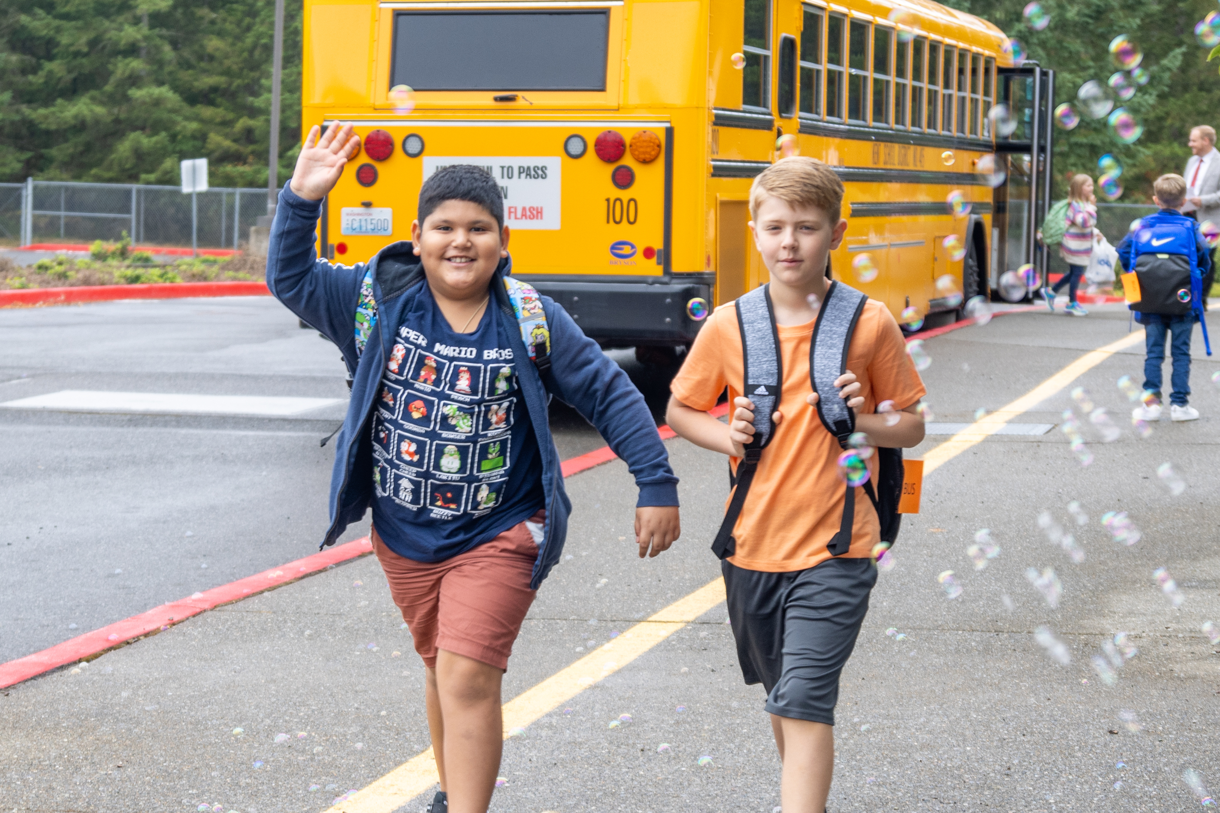 Two boys walking away from a yellow school bus, waving, with other children and a person standing near the bus in the background, surrounded by bubbles.
