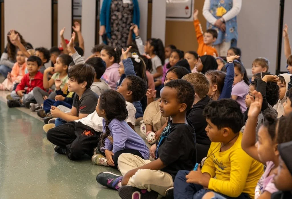 A large group of diverse children sitting cross-legged on the floor in a classroom or auditorium, many with hands raised, attentively participating in a discussion or activity.
