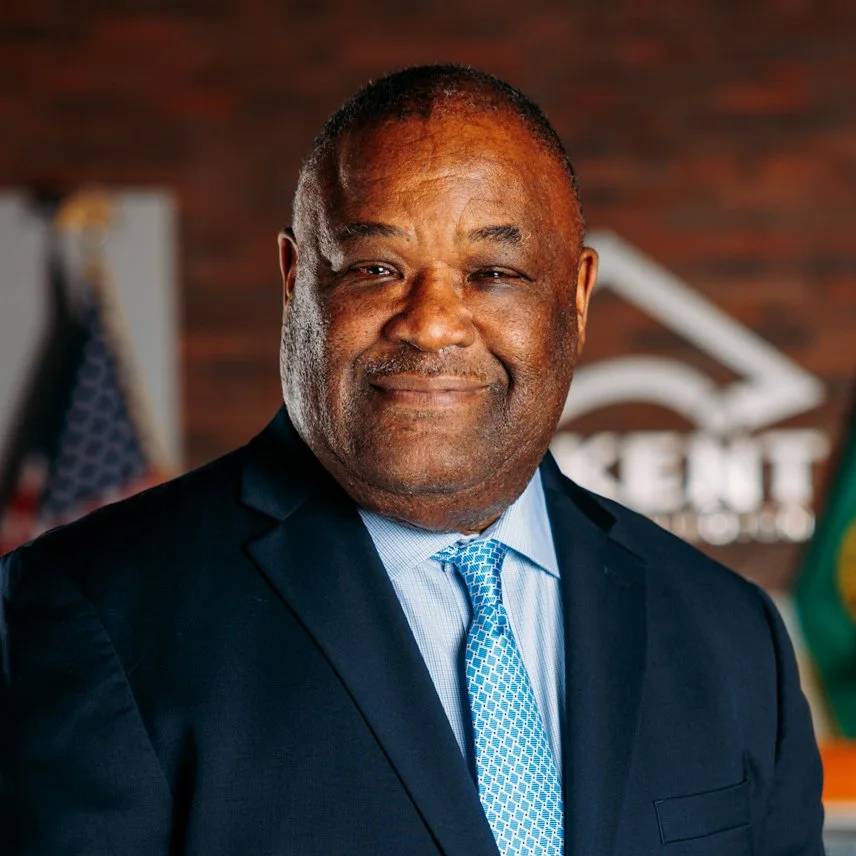 Portrait of a middle-aged man in a suit and tie, smiling, with a blurred office background and flags.