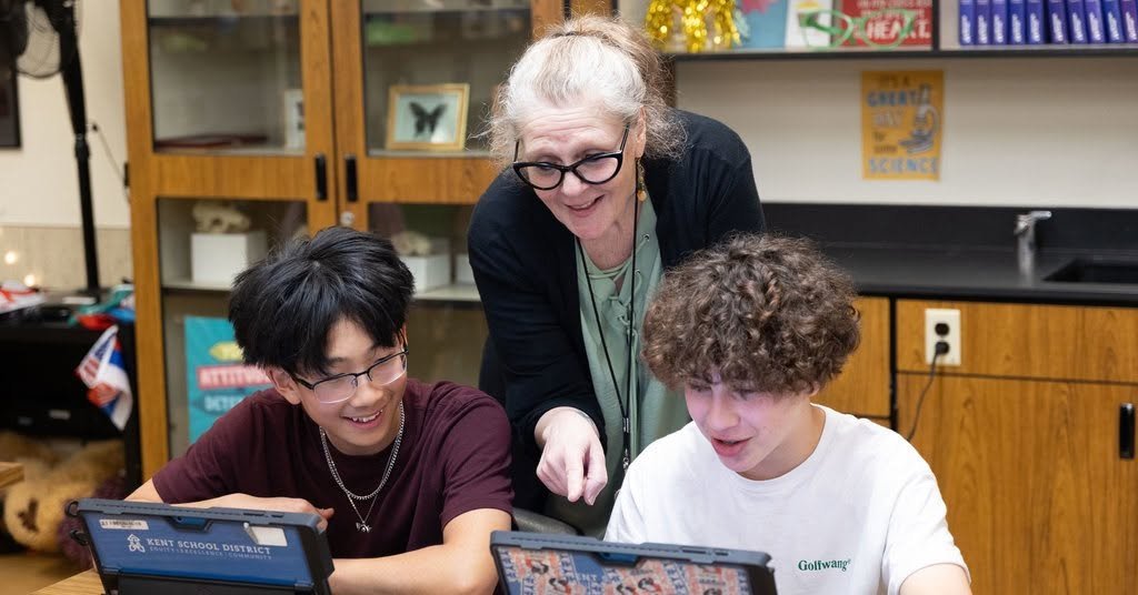 A teacher and two students in a classroom looking at tablets, smiling and engaged.