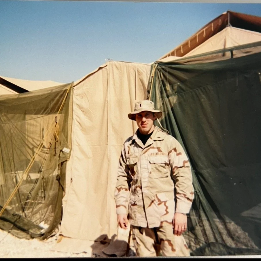 A soldier in desert camouflage uniform standing in front of military tents.