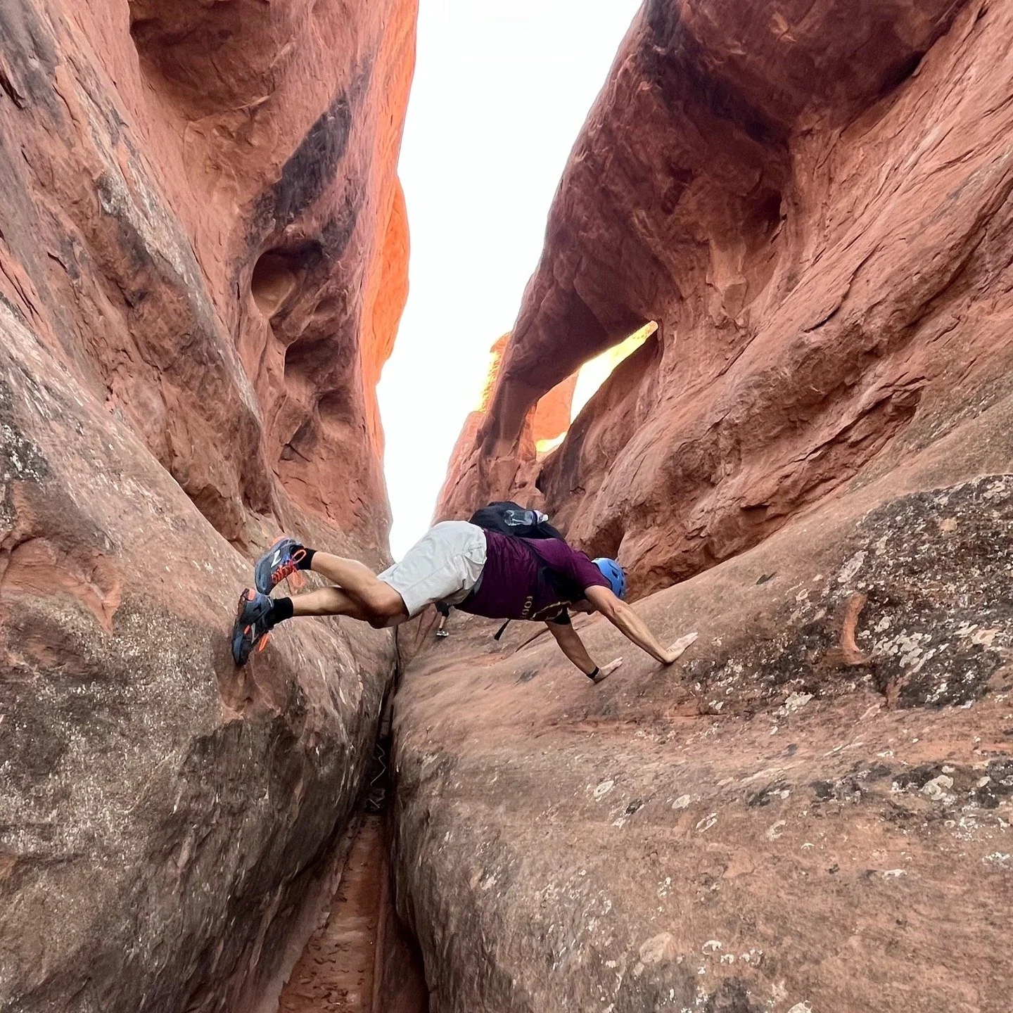 A person wearing a helmet and climbing gear is bouldering on a steep, narrow rock formation in a canyon with red and orange sandstone walls.