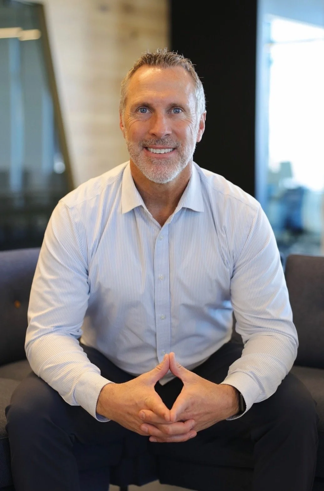 A smiling middle-aged man with gray hair and beard, wearing a light blue dress shirt, sitting on a dark gray sofa in a modern office or lounge area with large windows in the background.