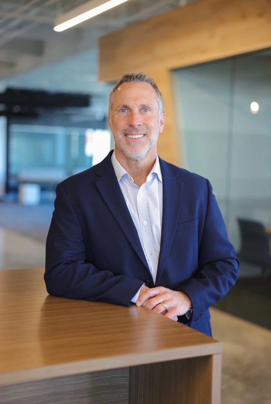 A middle-aged man with gray hair and a beard wearing a navy blue suit and white shirt, smiling and standing in a modern office environment with glass partitions and wood accents.