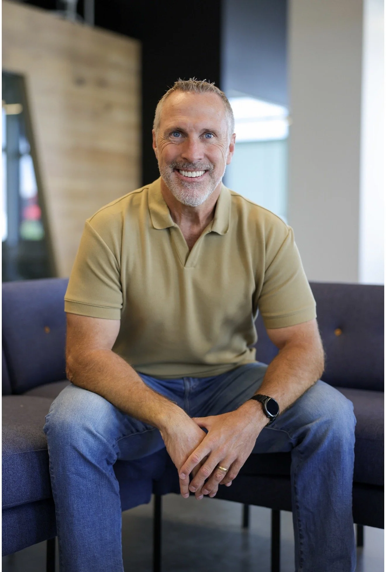 A smiling middle-aged man with gray hair and beard, wearing a tan polo shirt and jeans, sitting on a dark blue couch in a modern indoor setting with wooden and gray walls, and a window in the background.