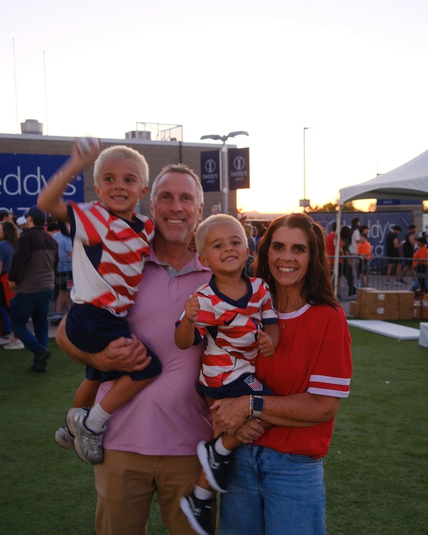 A multigenerational family at an outdoor event during sunset, with two young boys in football jerseys, and their parents holding them and smiling.