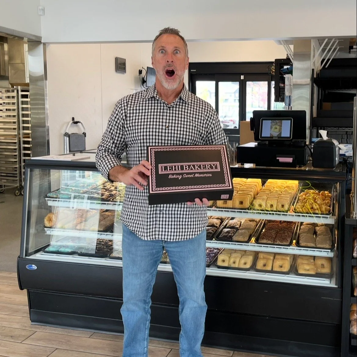 A man standing in front of a bakery display case holding a box labeled 'Lehi Bakery' with a surprised expression.
