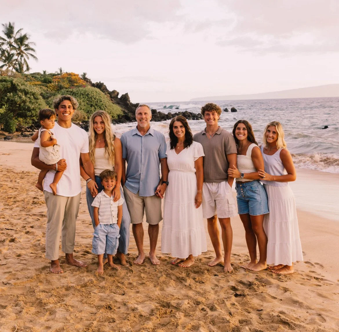 A diverse family standing on the beach during sunset, smiling, with ocean waves and greenery in the background.