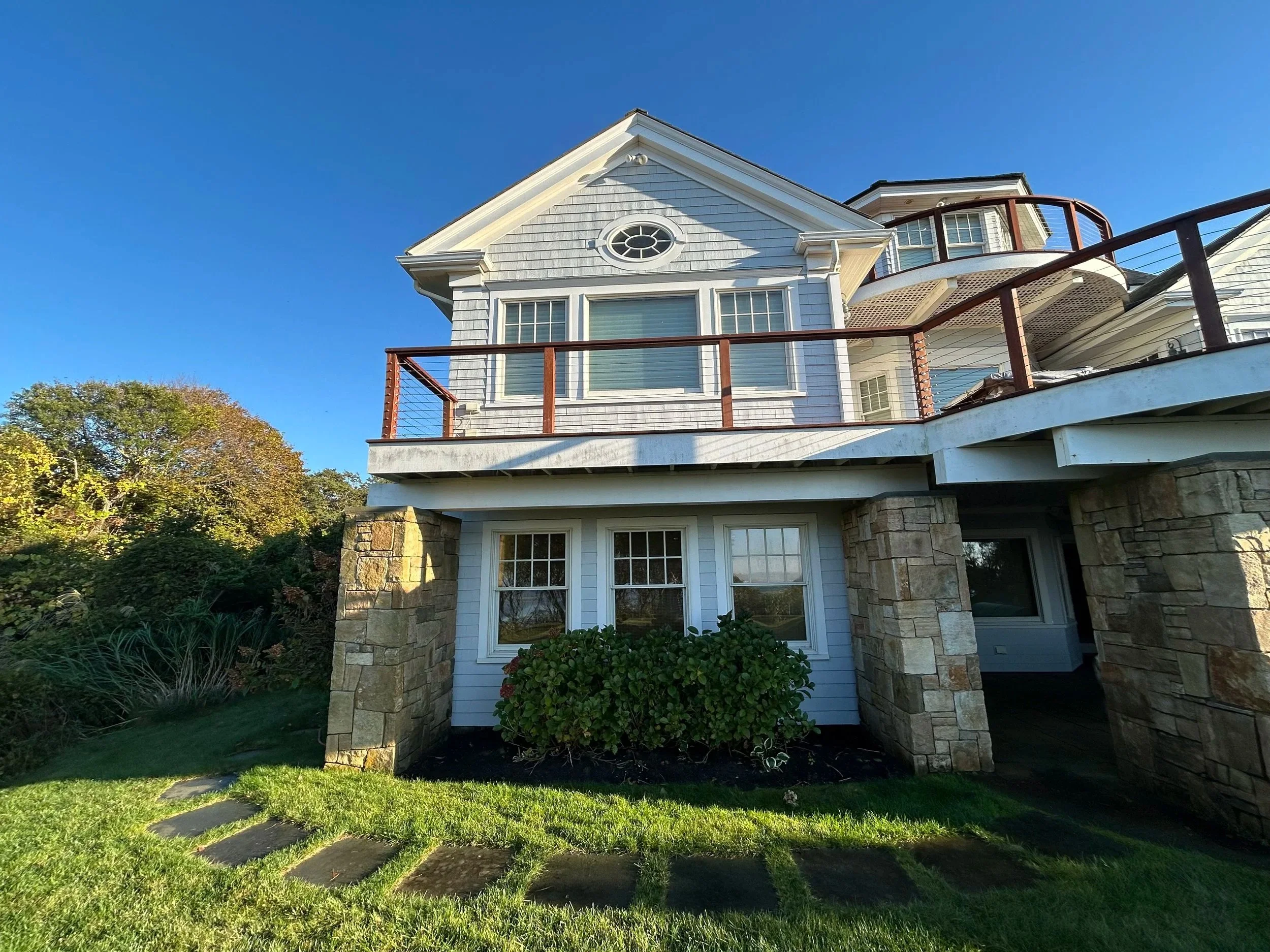 The image shows the back of a light blue house with a stone foundation, a balcony with a wooden railing, and multiple windows under a clear blue sky. There is a garden with green grass and a stone pathway in the front.