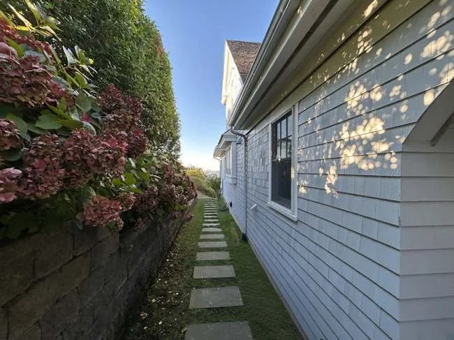 Side view of a house with white siding, a window, and a small pathway with square stepping stones, bordered by a flower bed with pink flowers on the left.