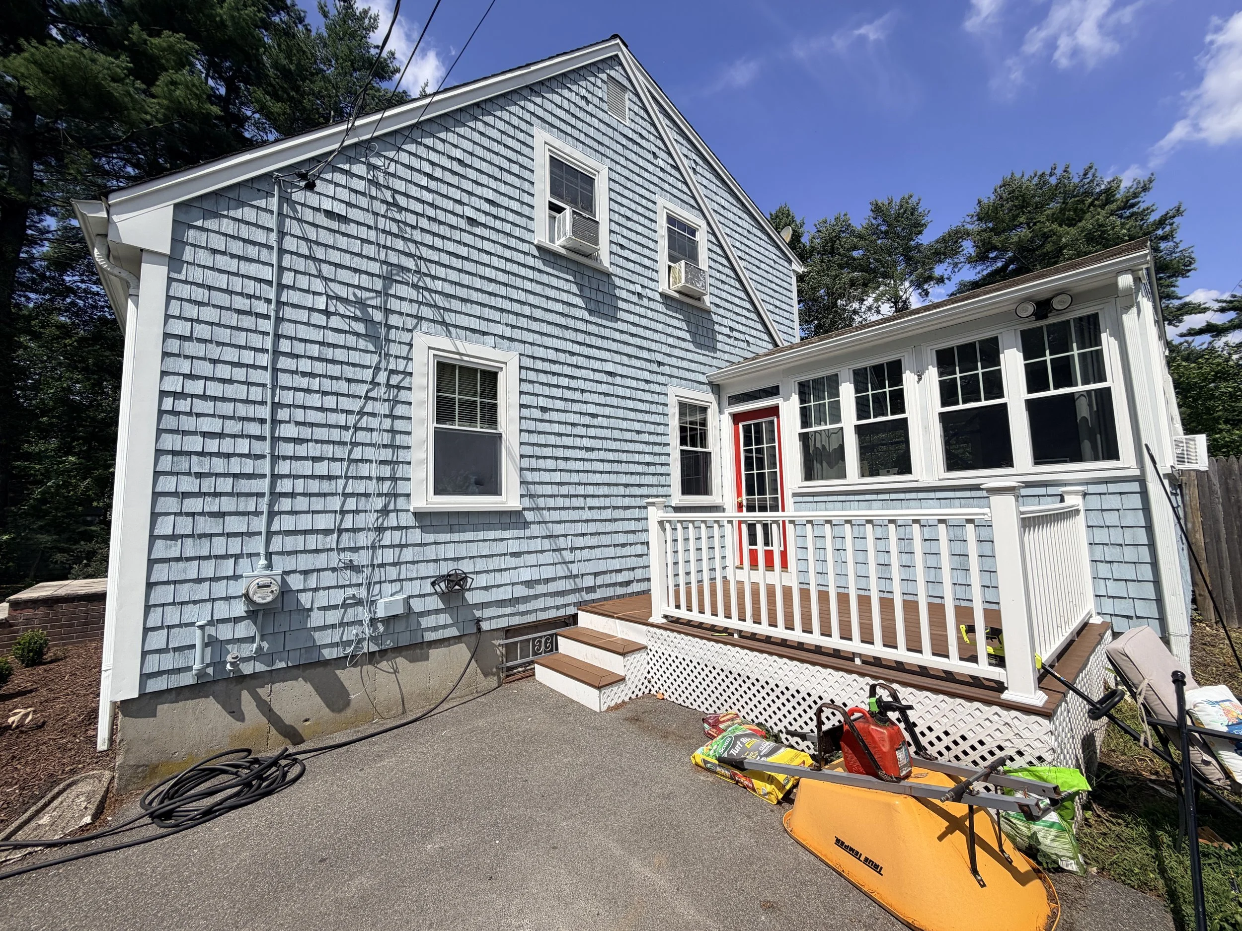 Rear of a two-story house with blue shingle siding, multiple windows with air conditioning units, and a small porch with wooden steps and a white railing. Items like garden supplies, tools, and outdoor equipment are scattered around the patio area. The sky is partly cloudy, and trees surround the house.