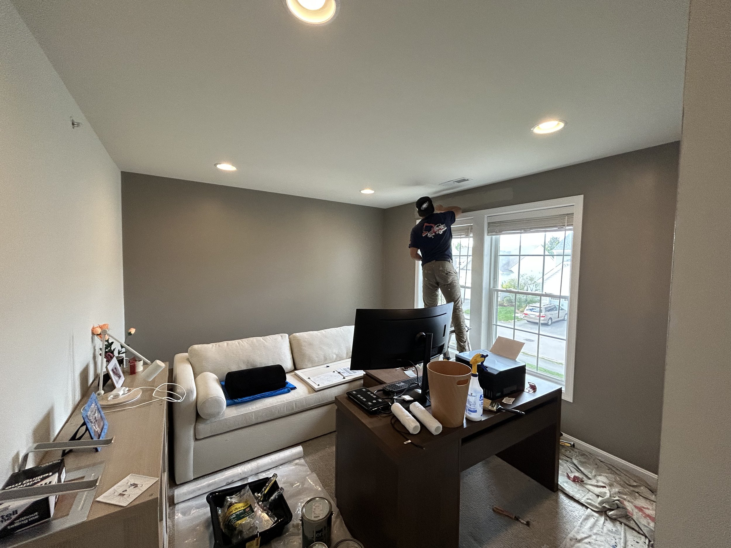 A man standing on a chair painting the ceiling of a living room with gray walls and a large window. The room has a white sofa, a table with various items, and clutter on the floor.