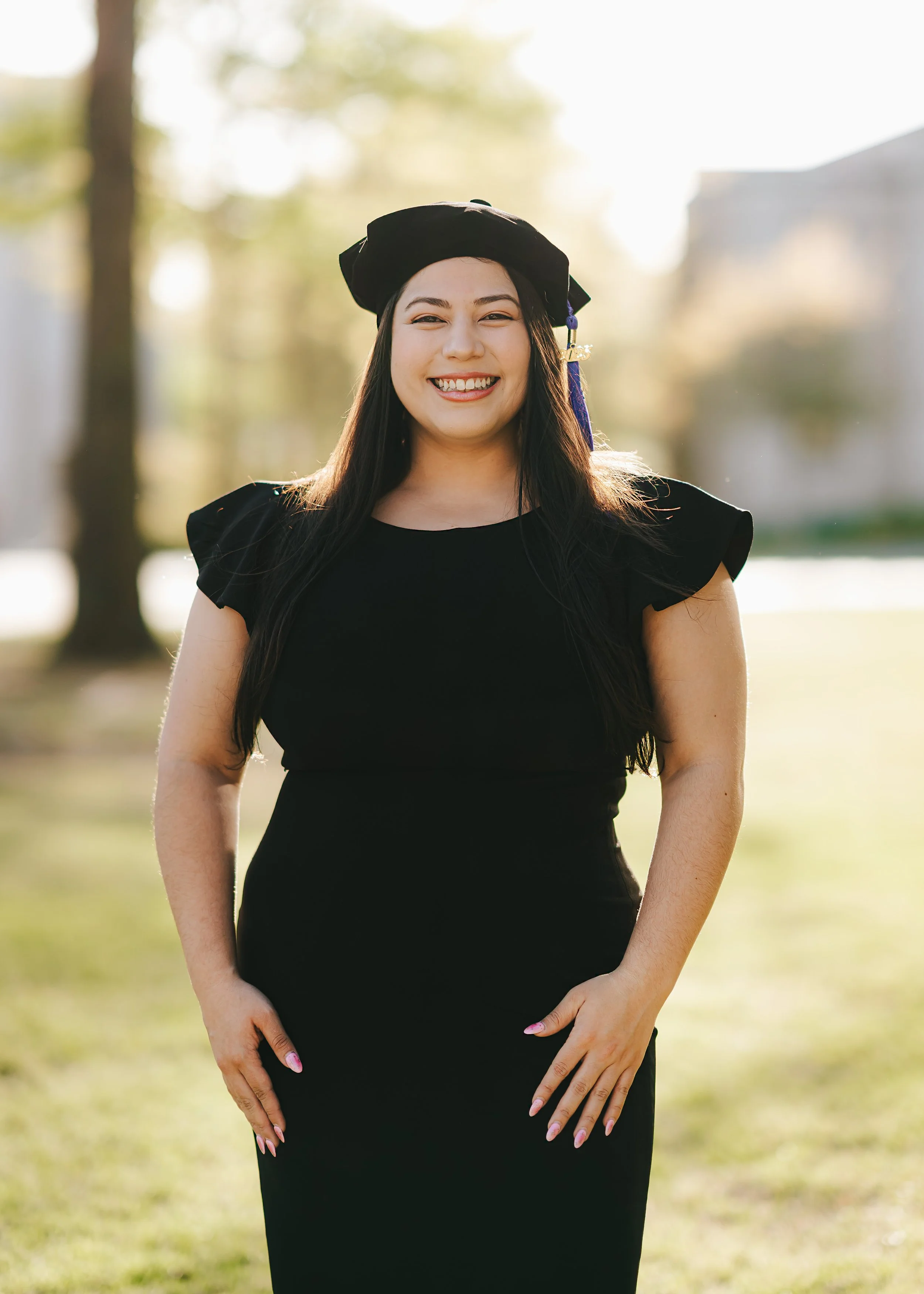 A young woman smiling in a black dress and graduation cap outdoors with trees and sunlight behind her.