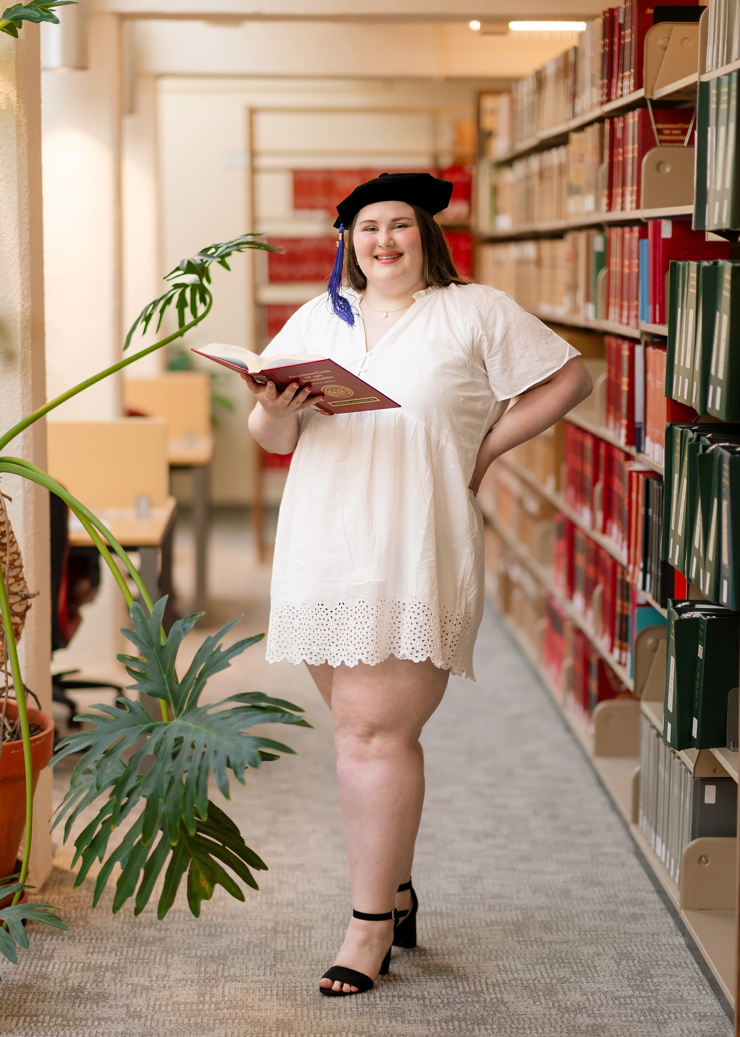 A woman in a white dress and black heels standing in a library, holding an open book, smiling.