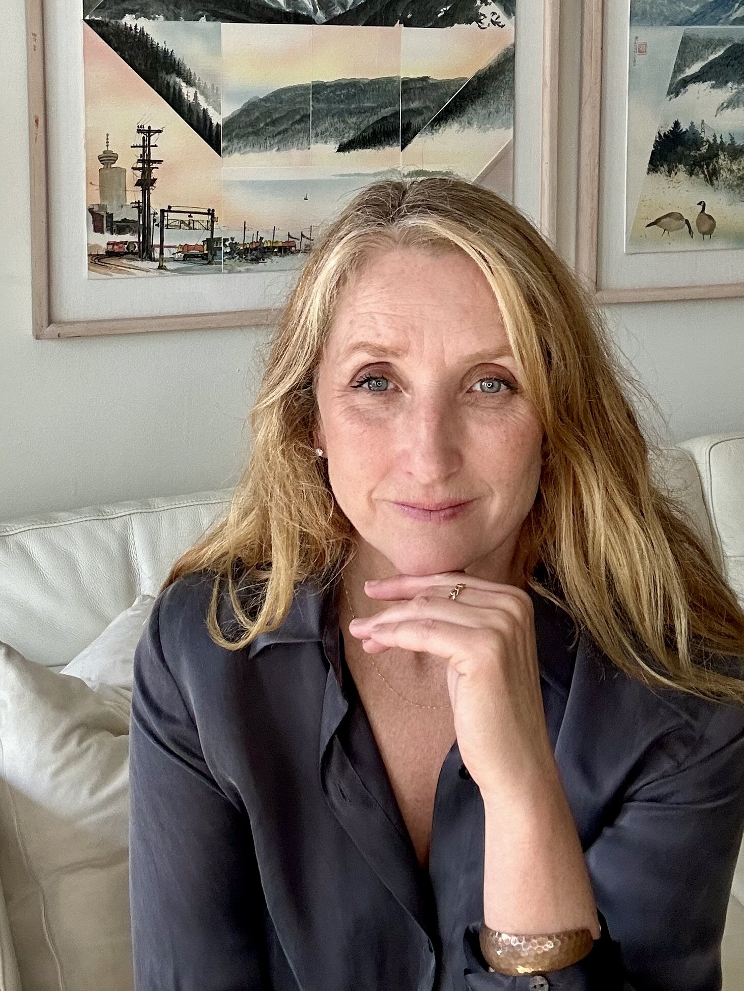 A woman with blonde hair and blue eyes sitting on a white couch in front of framed watercolor landscape paintings, resting her chin on her hand, wearing a dark shirt and gold jewelry.