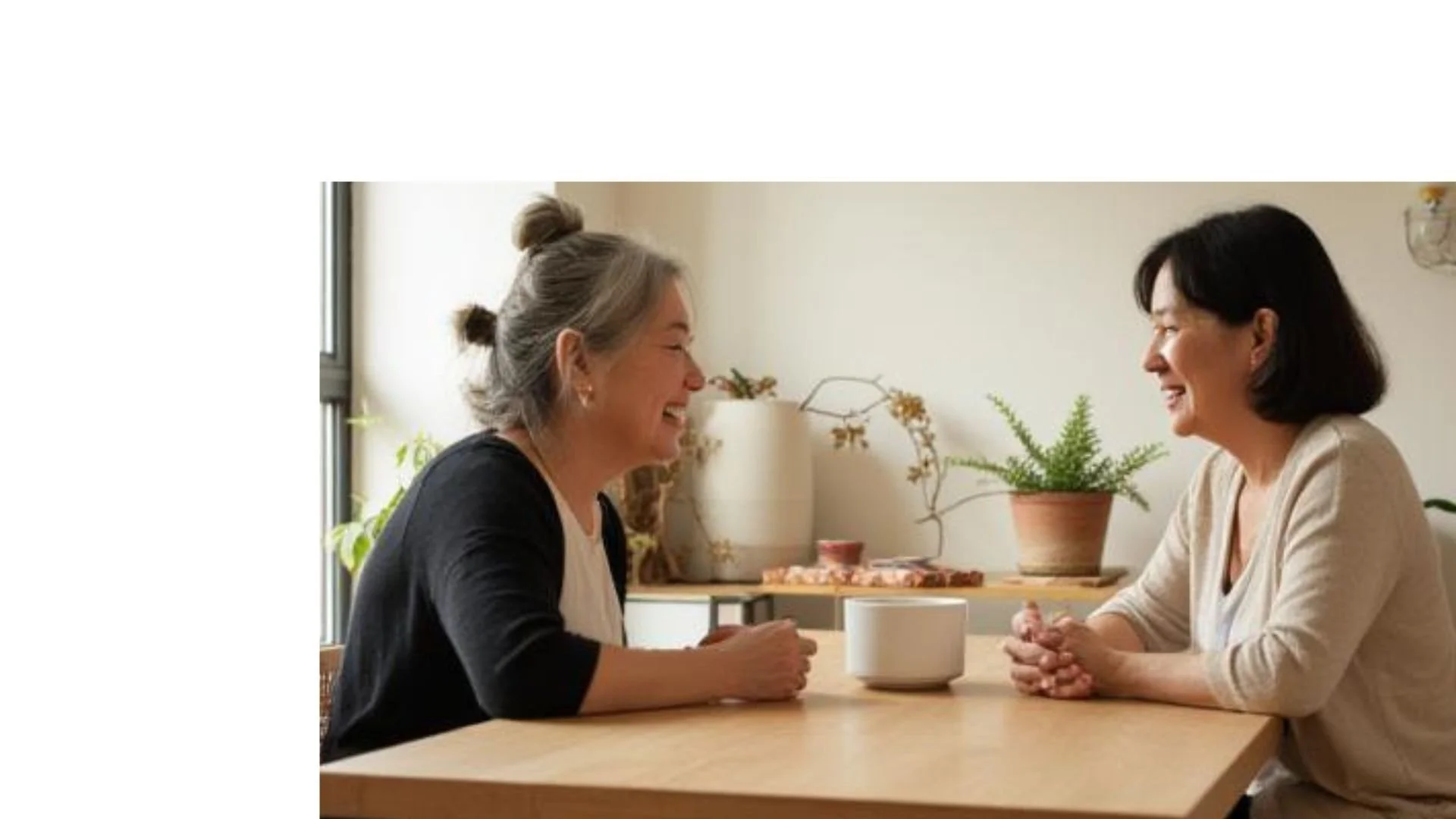 Two women are sitting at a wooden table facing each other, smiling and having a conversation, in a bright room with plants.
