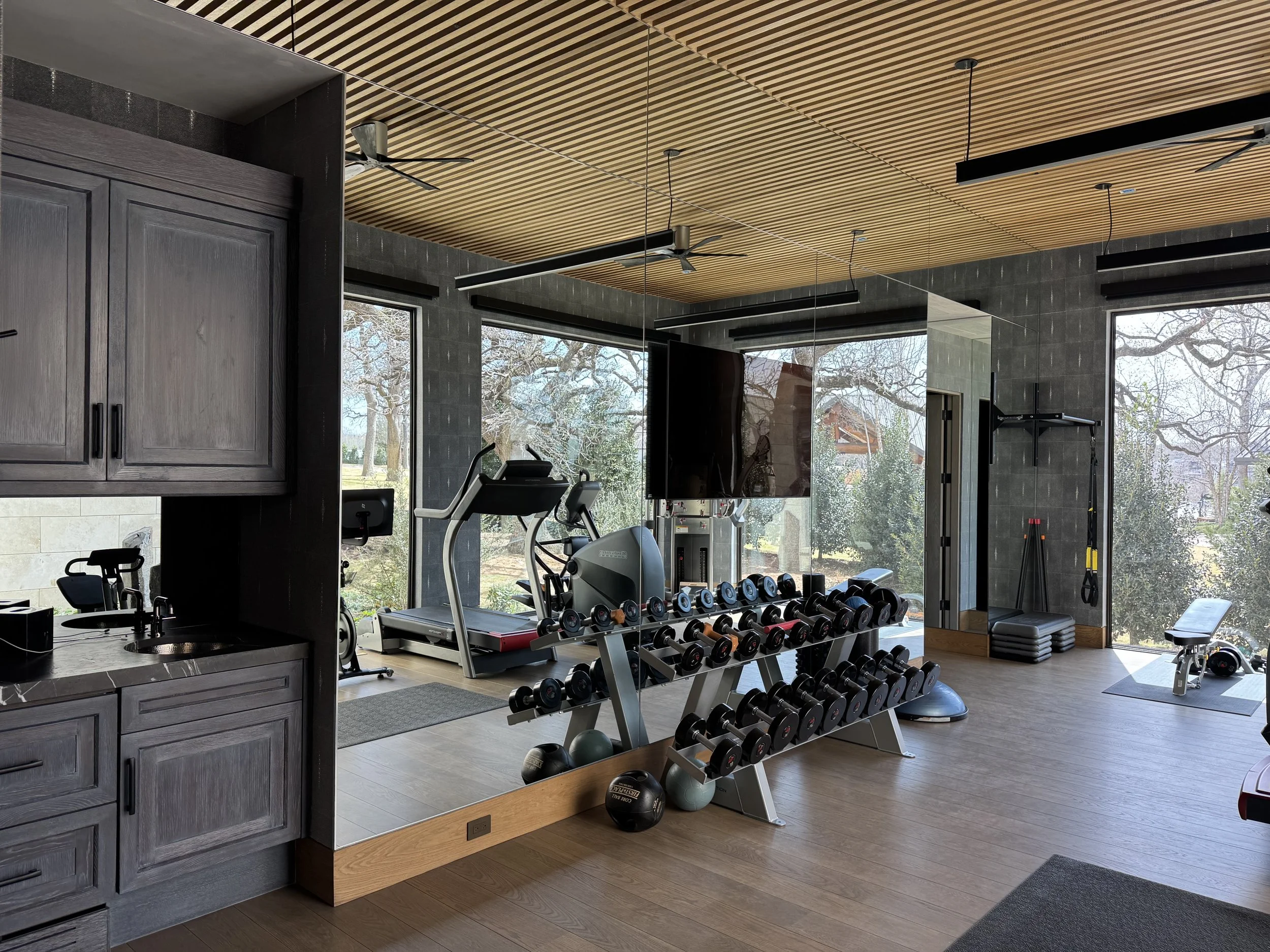 Interior of a modern gym with large windows, fitness equipment including treadmills, dumbbells, kettlebells, and exercise mats, and a wooden slatted ceiling.