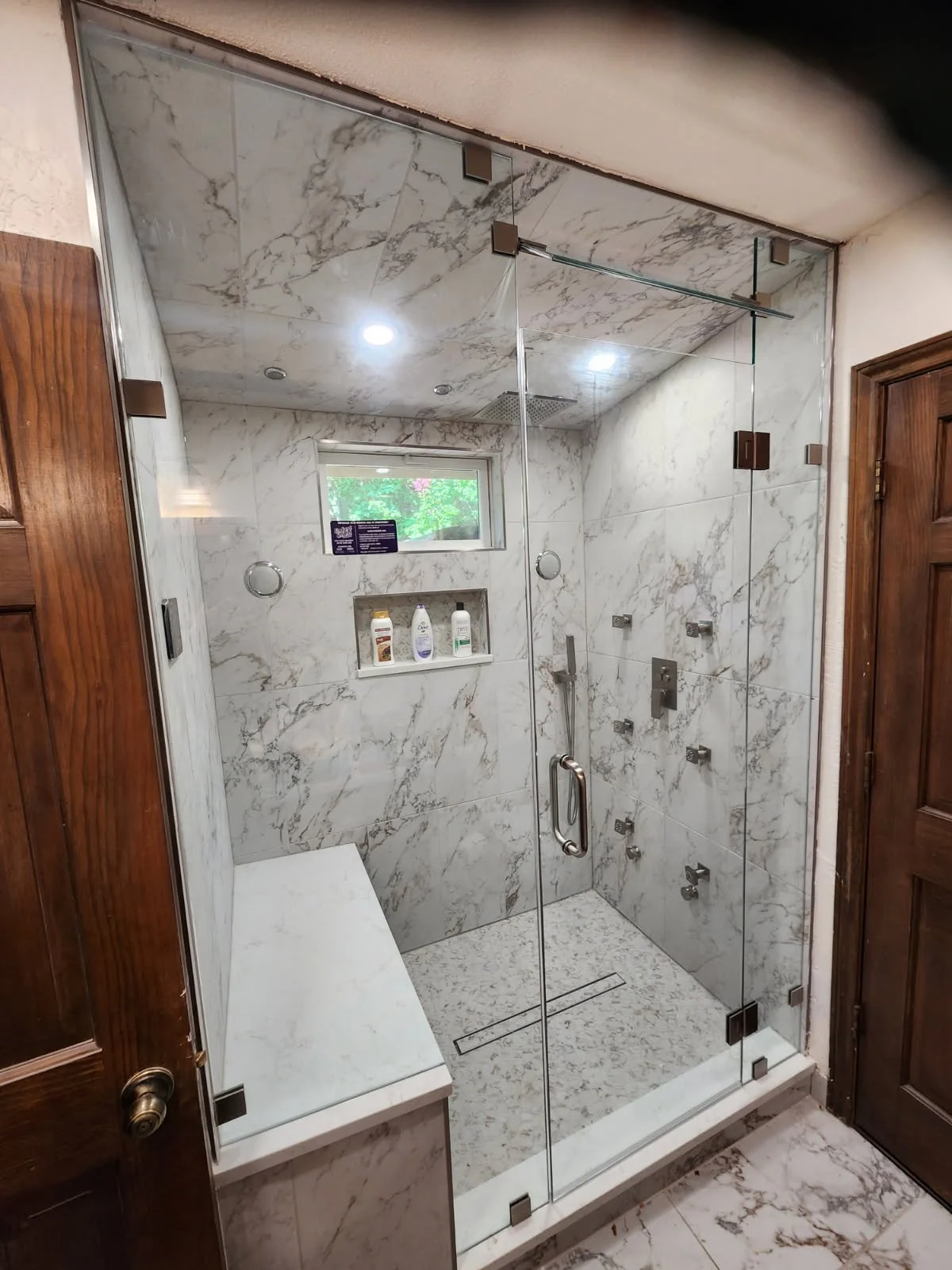 A modern walk-in shower with marble walls and ceiling, glass door, a small window, and built-in shelving with toiletries.