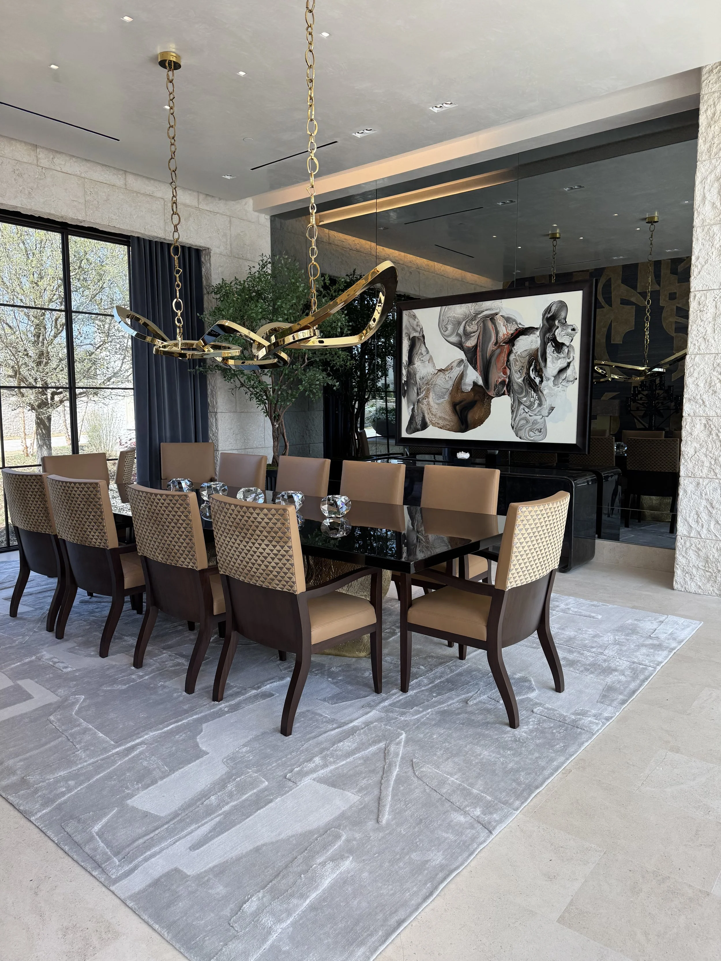 Formal dining room with a long black table, beige chairs, large window with dark curtains, modern chandelier, abstract artwork, indoor plant, stone and mirror decor, and a large light gray area rug.