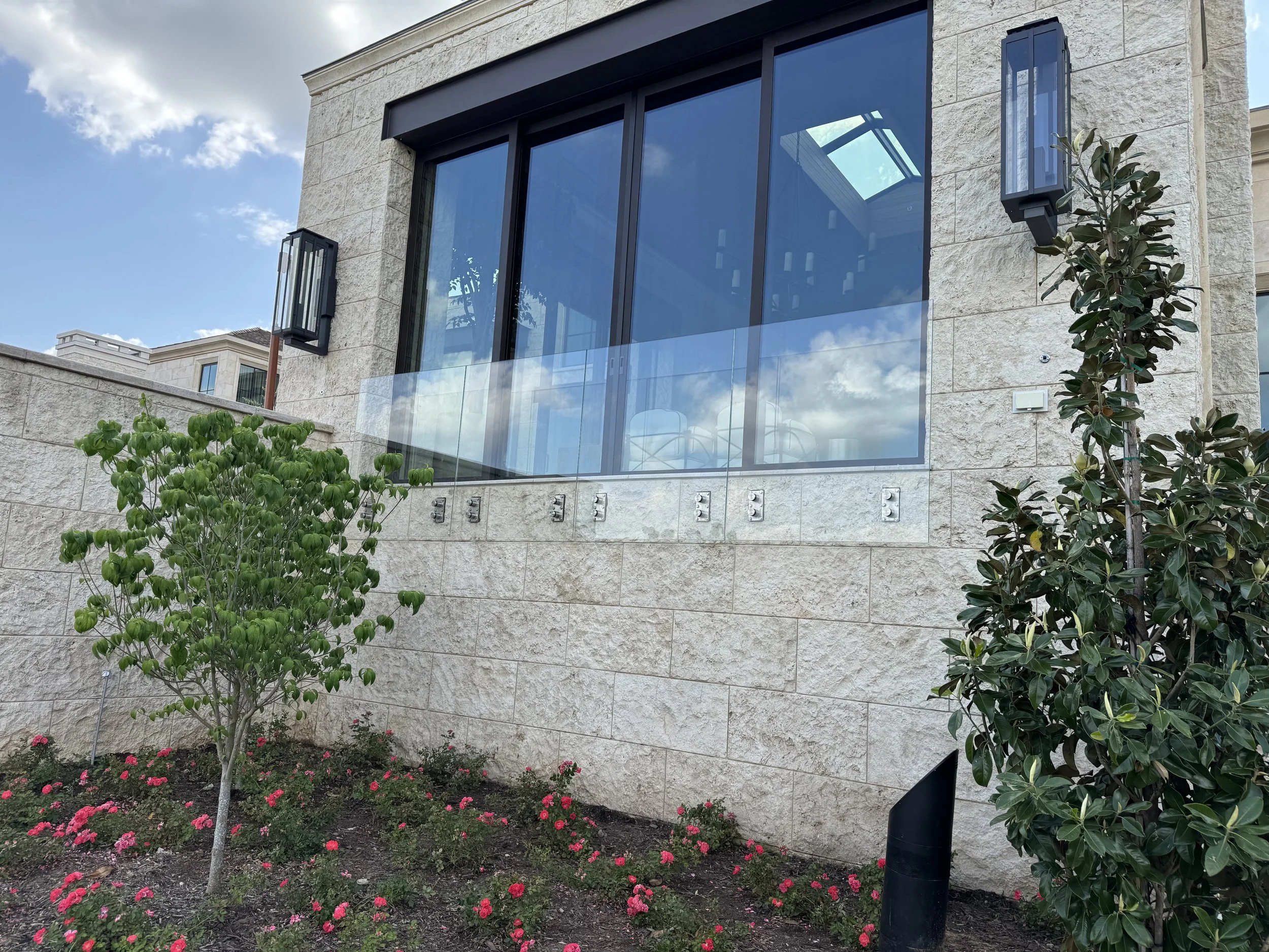 Exterior of a modern house with large glass windows, stone walls, outdoor lighting fixtures, and landscaped garden with trees and pink flowers.